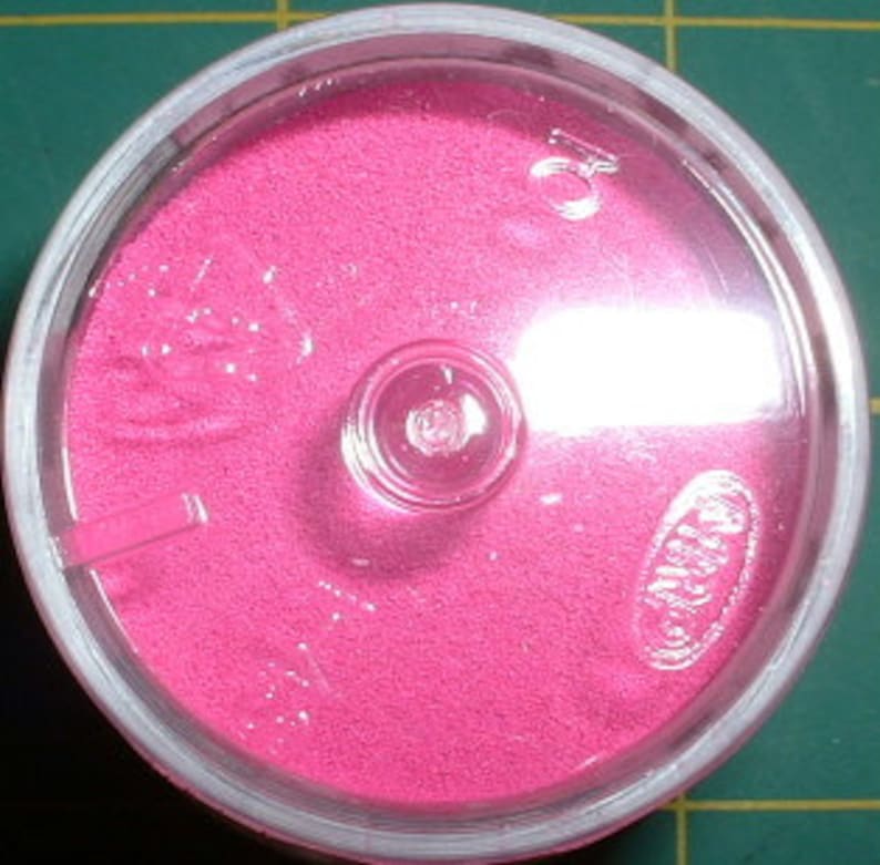 May include: A small, round container filled with bright pink sand. The container has a clear lid with a small hole in the center.