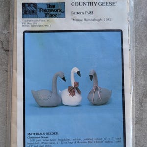 May include: A sewing pattern packet titled Country Geese Pattern P-22 by Marine Bumbalough, 1982. The cover features three fabric geese: two grey and one white, with the white goose wearing a red plaid ribbon and one grey goose a pink ribbon.