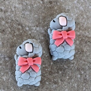 May include: Two gray sleeping sheep with pink bows. The sheep are made of polymer clay and have a textured surface.