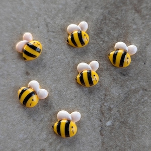 May include: Six small bee-shaped buttons, each featuring a yellow body with black horizontal stripes and two white wings. The buttons have a smooth, glossy finish and are arranged in a scattered pattern, showcasing the insect-inspired design.