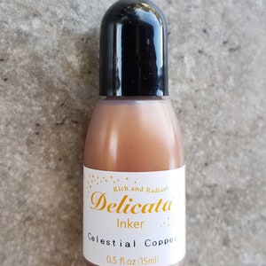 Op de afbeelding: Een klein flesje Celestial Copper inkt van Delicata Inker. De fles is lichtbruin van kleur met een zwarte dop. Het etiket is wit met gouden letters en vermeldt "Rich and Radiant Delicata Inker Celestial Copper 0.5 fl.oz (15ml)"