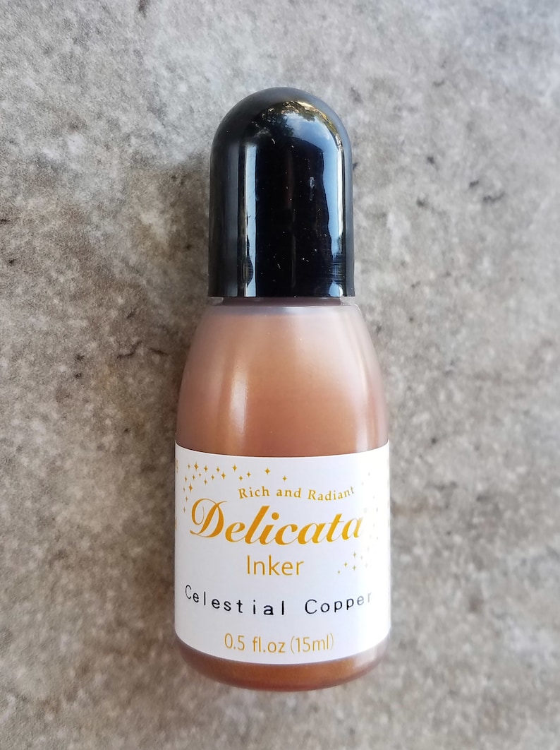 Op de afbeelding: Een klein flesje Celestial Copper inkt van Delicata Inker. De fles is lichtbruin van kleur met een zwarte dop. Het etiket is wit met gouden letters en vermeldt "Rich and Radiant Delicata Inker Celestial Copper 0.5 fl.oz (15ml)"