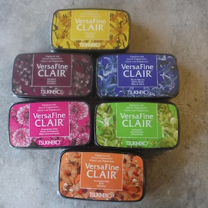 May include: Seven different colored ink pads with floral designs and the text "VersaFine CLAIR" on each. The ink pads are made by Tsukineko.