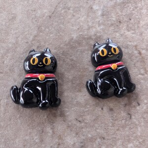 May include: Two small, glossy black cat figurines in a seated position. Each cat features large, bright yellow eyes, a small smile, and a red collar with a tiny yellow bell detail. The figures have a smooth, reflective finish and are identical.