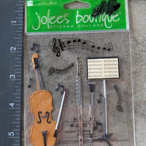 May include: A clear package of Jolee's Boutique sticker collage. The stickers feature musical instruments like a violin, flute, and clarinet, along with musical notes and sheet music. The violin is brown and the other instruments are silver.