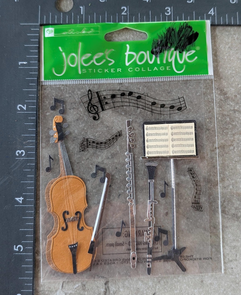May include: A clear package of Jolee's Boutique sticker collage. The stickers feature musical instruments like a violin, flute, and clarinet, along with musical notes and sheet music. The violin is brown and the other instruments are silver.