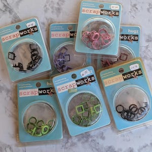 May include: Assortment of six packages of scrapbooking embellishments. Each package contains a variety of small metal shapes in different colors, including pink, purple, green, and black. The packages are labeled "Scrapworks" and feature a variety of designs.