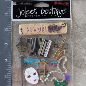 May include: A sticker collage by Jolee's Boutique featuring New Orleans themed designs. Includes a wooden sign with "New Orleans" text, an accordion, saxophone, mask, and beads. The collage is in a clear plastic package.