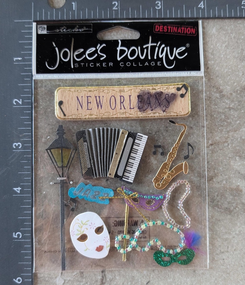 May include: A sticker collage by Jolee's Boutique featuring New Orleans themed designs. Includes a wooden sign with "New Orleans" text, an accordion, saxophone, mask, and beads. The collage is in a clear plastic package.
