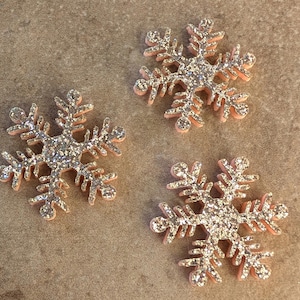 May include: Three pink glitter snowflake ornaments. The snowflakes are made of foam and have a silver glitter finish.