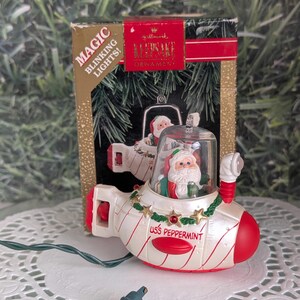 May include: A Hallmark Christmas ornament shaped like a submarine, with Santa Claus inside. The ornament is white with red accents and the words "USS Peppermint." The box reads "Magic Blinking Lights!"