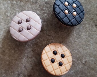May include: Three round buttons with a grid pattern. The buttons are in different colors: black, light pink, and light orange. Each button has four small round accents in the center.