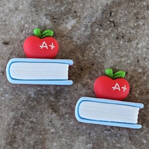 May include: Two decorative school-themed embellishments. Each features a red apple with a white "A+" and a green stem, resting on a light blue book with white pages. The items are designed for crafts or scrapbooking.