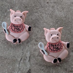 May include: Two pink pig figurines with black hooves and ears. Each pig wears a bib with the words "Feed Me" and holds a white spoon. The figurines are sitting on a gray surface.