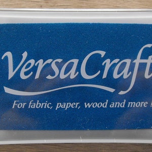 May include: A clear plastic container with a blue ink pad inside. The ink pad has the text "VersaCraft&trade;" in white lettering and the text "For fabric, paper, wood and more!" in white lettering.