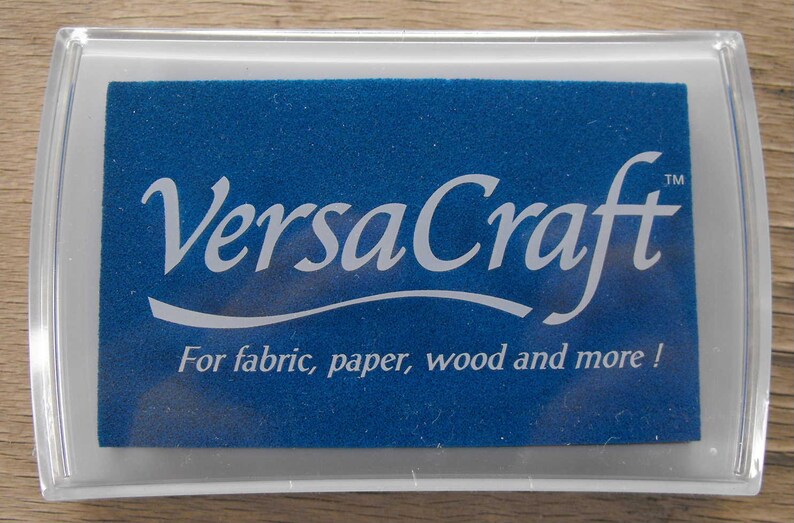 May include: A clear plastic container with a blue ink pad inside. The ink pad has the text "VersaCraft&trade;" in white lettering and the text "For fabric, paper, wood and more!" in white lettering.