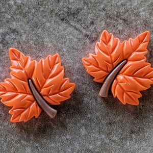 May include: Two orange maple leaf-shaped decorative items. Each leaf has detailed veining and a brown stem. The leaves are a vibrant orange color, suggesting an autumn theme. The items are likely decorative accessories or embellishments.