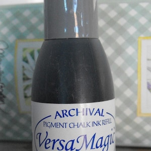 May include: A black ink bottle with a gray cap. The label reads "Archival Pigment Chalk Ink Refill VersaMagic 91 Midnight Black".