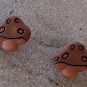 May include: Two brown mushroom-shaped decorative buttons with a light brown stem and dark brown details. The buttons have a rounded top with circular indentations. The buttons are placed on a textured, light gray surface.