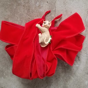 May include: A red velvet ribbon with a small, beige figurine of a mouse wearing a green holly sprig.