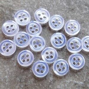 May include: A collection of 14 clear plastic buttons with four holes each. The buttons are arranged in a circle on a gray surface.