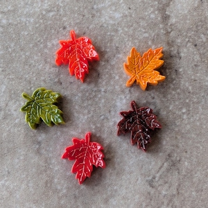 May include: Five small maple leaf-shaped ornaments in various colors. The set includes one bright red leaf, one orange leaf, one dark brown leaf, one olive green leaf, and one lime green leaf. Each piece features detailed embossed veins and a glossy finish.