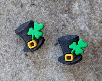Puede incluir: Dos dijes de sombrero de copa negro, cada uno con un trébol verde brillante de tres hojas en el lateral y un detalle de hebilla cuadrada amarilla en la banda del sombrero. Estos pequeños artículos decorativos tienen forma de mini sombreros.