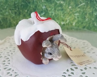 May include: A ceramic ornament shaped like a red apple with white frosting. A small gray rabbit peeks out of a bite taken out of the apple. The ornament has a red string attached to the top and a tag that reads "Silvestri Corp. Made in Taiwan."