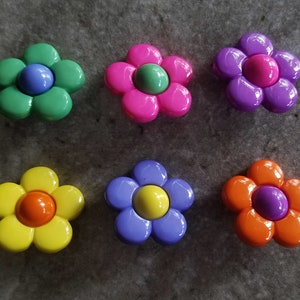 May include: Six plastic flower-shaped buttons arranged in two rows. The top row features green with blue center, pink with green center, and purple with pink center. The bottom row has yellow with orange center, blue with yellow center, and orange with purple center.