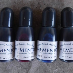 Puede incluir: Cuatro botellas peque&ntilde;as de tinta Memento resistente a la decoloraci&oacute;n en varios colores: Grape Jelly, Lilac Posies, Bahama Blue y Nautical Blue. Cada botella tiene una tapa gris oscuro y una etiqueta blanca con texto negro. La tinta es a base de agua y sin &aacute;cido.