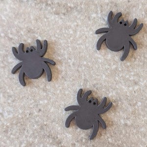 May include: Three dark gray spider-shaped embellishments with curved legs and small eye holes. The spiders are arranged on a light tan, textured surface. These decorative elements are suitable for craft projects.