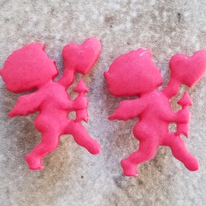 May include: Two pink cherub figurines holding hearts. The cherubs are made of a smooth, plastic-like material and have wings and arrows.