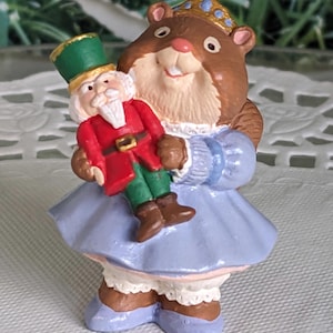 May include: A brown and white figurine of a squirrel wearing a blue dress and a gold crown, holding a green and red nutcracker figurine.
