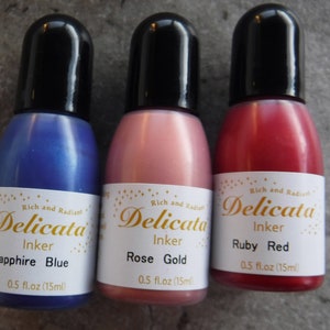 Op de afbeelding: Drie kleine flesjes Delicata-inkt in saffierblauw, roosgoud en robijnrood. Elk flesje is voorzien van het Delicata-logo en de inktkleur. De flesjes zijn elk 0,5 fl.oz (15ml).
