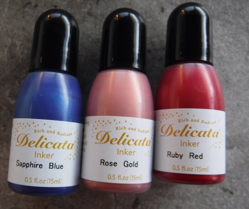 Op de afbeelding: Drie kleine flesjes Delicata-inkt in saffierblauw, roosgoud en robijnrood. Elk flesje is voorzien van het Delicata-logo en de inktkleur. De flesjes zijn elk 0,5 fl.oz (15ml).