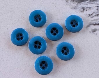 May include: Seven round, blue buttons with four holes each are scattered on a light-colored surface. The buttons have a slightly marbled appearance, and the color is a medium shade of blue.