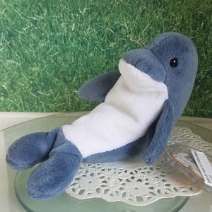 May include: A blue and white plush dolphin toy. The dolphin is lying on its side with its head turned to the side. The dolphin has a white belly and a blue back. The toy has a Ty tag attached to it.