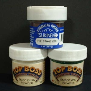 May include: Three containers of embossing powder. The container on the left is labeled "Top Boss Embossing Powder" and is a light beige color. The container in the middle is labeled "Tsukineko Embossing Powder #12 Stone Red" and is a blue and white container with a red powder inside. The container on the right is labeled "Top Boss Embossing Powder" and is a green color.