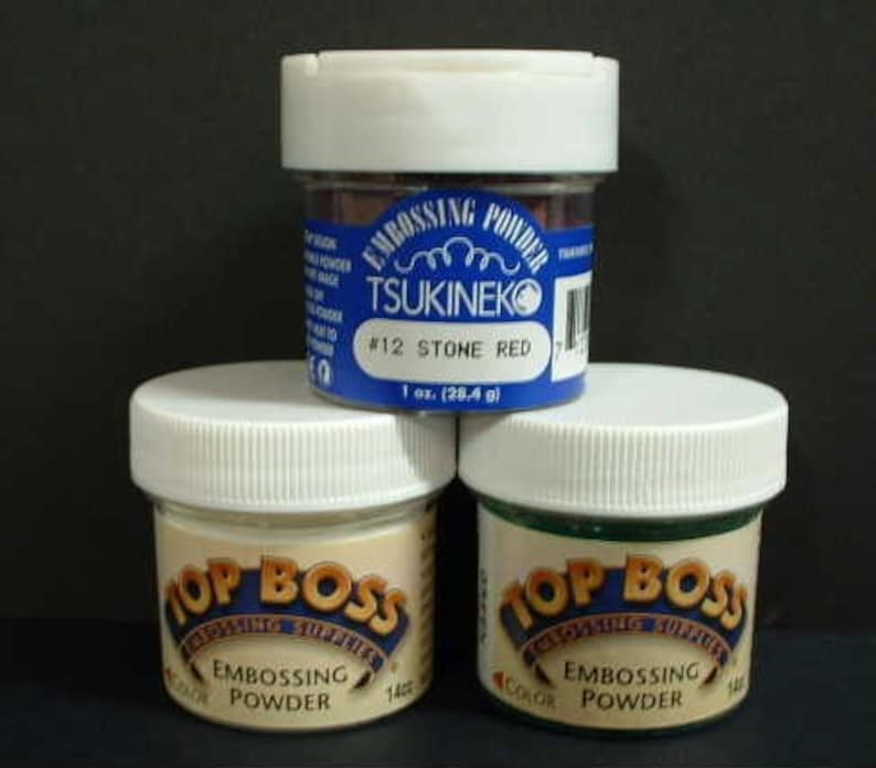 May include: Three containers of embossing powder. The container on the left is labeled "Top Boss Embossing Powder" and is a light beige color. The container in the middle is labeled "Tsukineko Embossing Powder #12 Stone Red" and is a blue and white container with a red powder inside. The container on the right is labeled "Top Boss Embossing Powder" and is a green color.
