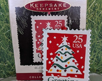 May include: A white metal Christmas ornament shaped like a postage stamp with a red background and a green and white Christmas tree with red and yellow ornaments. The stamp says "25 USA" and "Greetings".