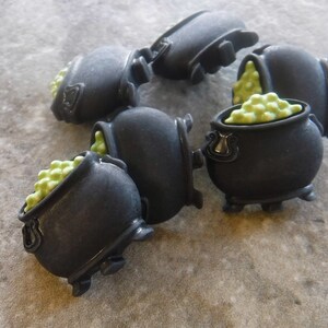 May include: A collection of black cauldron-shaped embellishments, each filled with light green, round, bubbly objects. The cauldrons have small legs and handles. The image is taken from a slightly elevated angle, showcasing the details of the miniature cauldrons.