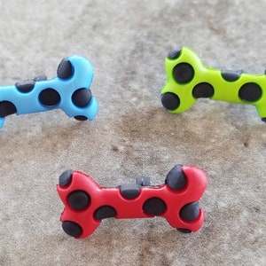 May include: Three bone-shaped charms in bright blue, lime green, and red. Each charm features a smooth, solid-colored surface with multiple circular black indentations distributed across the front, creating a distinct textured appearance.