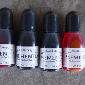 Puede incluir: Cuatro botellas peque&ntilde;as de tinta de tinte resistente a la decoloraci&oacute;n Memento en varios colores. Las botellas tienen tapas negras y etiquetas blancas con el nombre de la marca y los nombres de los colores: Lady Bug, Potter's Clay, Teal Zeal y Dandelion. La tinta es a base de agua y sin &aacute;cido.