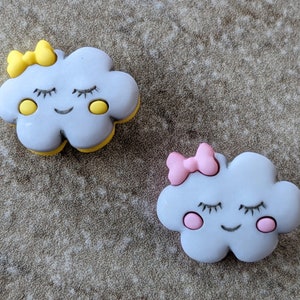 May include: Two gray felt cloud-shaped buttons with smiling faces. One cloud has a yellow bow and yellow eyes, the other has a pink bow and pink cheeks.
