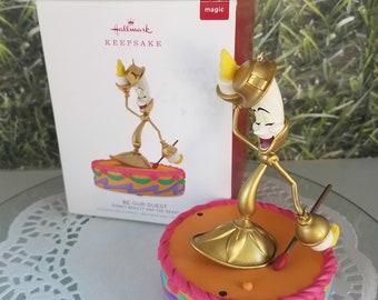 May include: A gold-colored musical ornament of Lumiere, the animated candlestick character from Disney's Beauty and the Beast. The ornament is on a cake-shaped base with a pink, orange, and green design. The ornament plays the song "Be Our Guest".