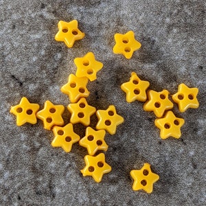 May include: A collection of 14 yellow star-shaped buttons with five points. The buttons are arranged in a loose, asymmetrical pattern.