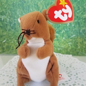 May include: A brown plush squirrel toy with a white belly and a red heart-shaped tag. The tag has the word "ty" in white and a gold star. The squirrel has a small black nose and whiskers.