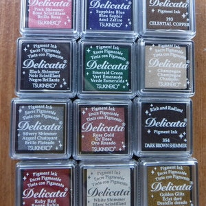 May include: Twelve small, square, clear plastic containers with black ink pads inside. Each container has a label with the brand name "Delicata" and the ink color name. The ink colors include: Pink Shimmer, Sapphire Blue, Celestial Copper, Black Shimmer, Emerald Green, Champagne, Silvery Shimmer, Rose Gold, Dark Brown Shimmer, Ruby Red, White Shimmer, and Golden Glitz.