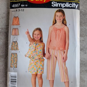 May include: Simplicity sewing pattern for girls' clothing, size A 3-12. The pattern features illustrations of tops and shorts. The package has the text "IT'S SO easy" and "Simplicity". The girls in the image are wearing the clothing.