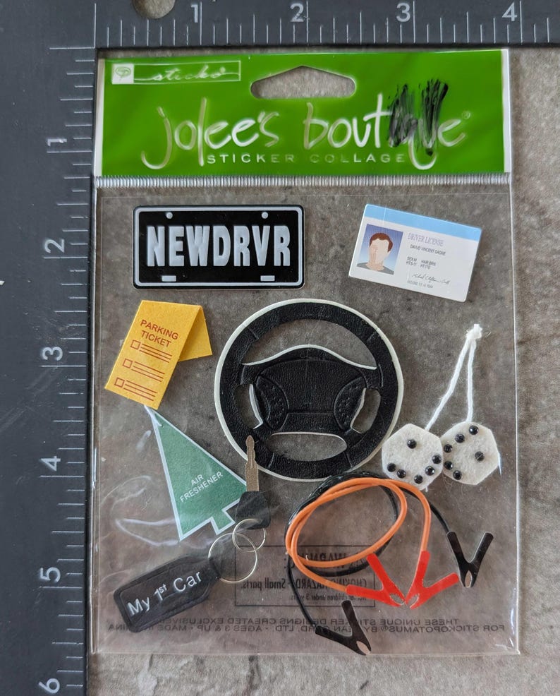 May include: A sticker collage featuring car-themed elements. Includes a license plate that reads "NEWDRVR", a steering wheel, a parking ticket, a driver's license, a key, dice, jumper cables, and an air freshener. The text "My 1st Car" is also visible.